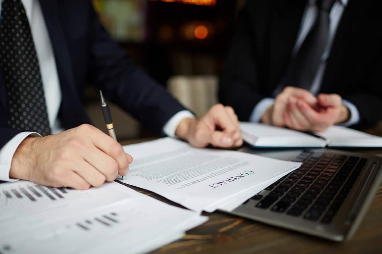 Two people in business suits review a contract document next to a laptop, using a pen for pointing.