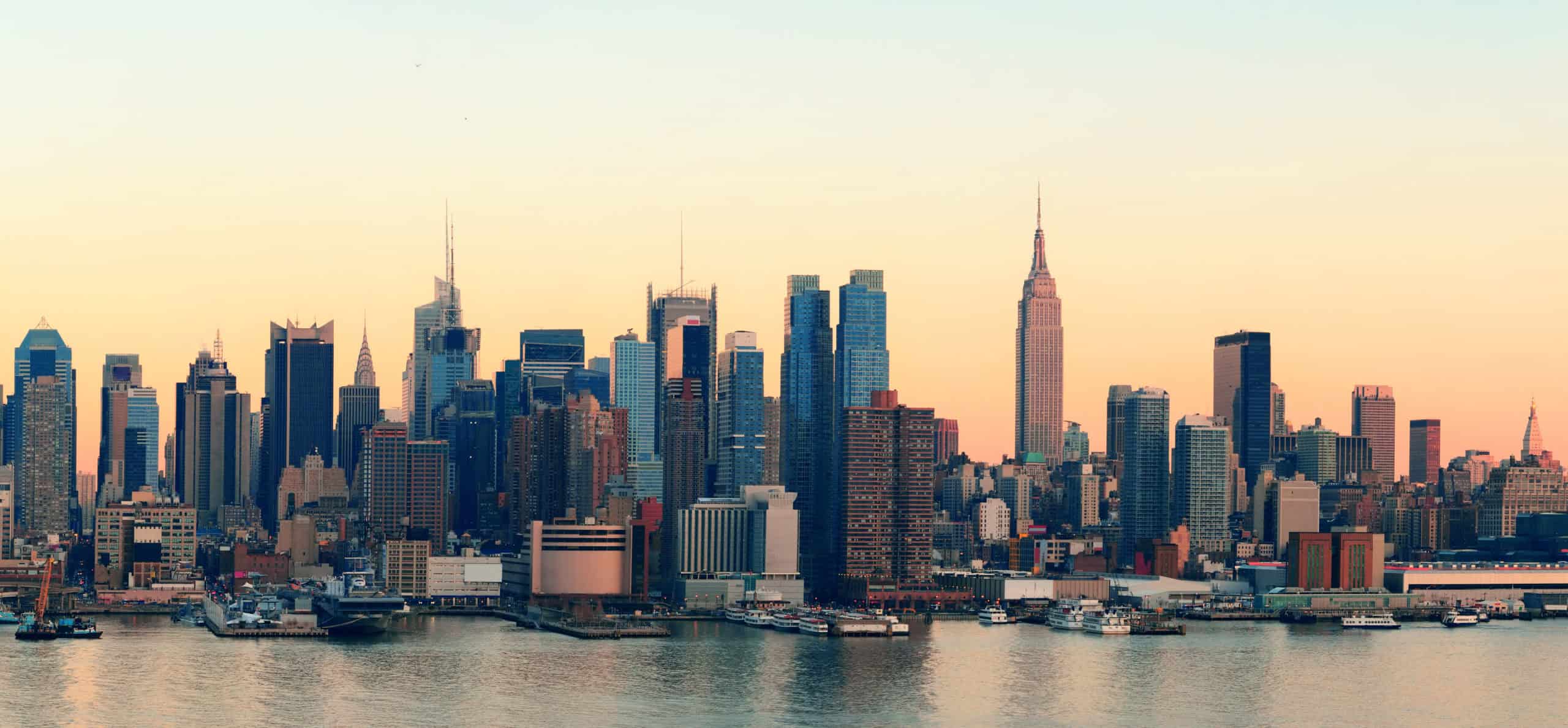 Skyline of a city with numerous skyscrapers and buildings, including a prominent tower, at dusk. A body of water is visible in the foreground.