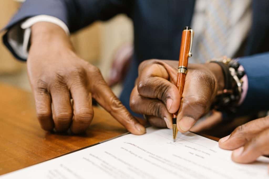 A person signs a document with a pen while another person points at the signature line on a wooden table.