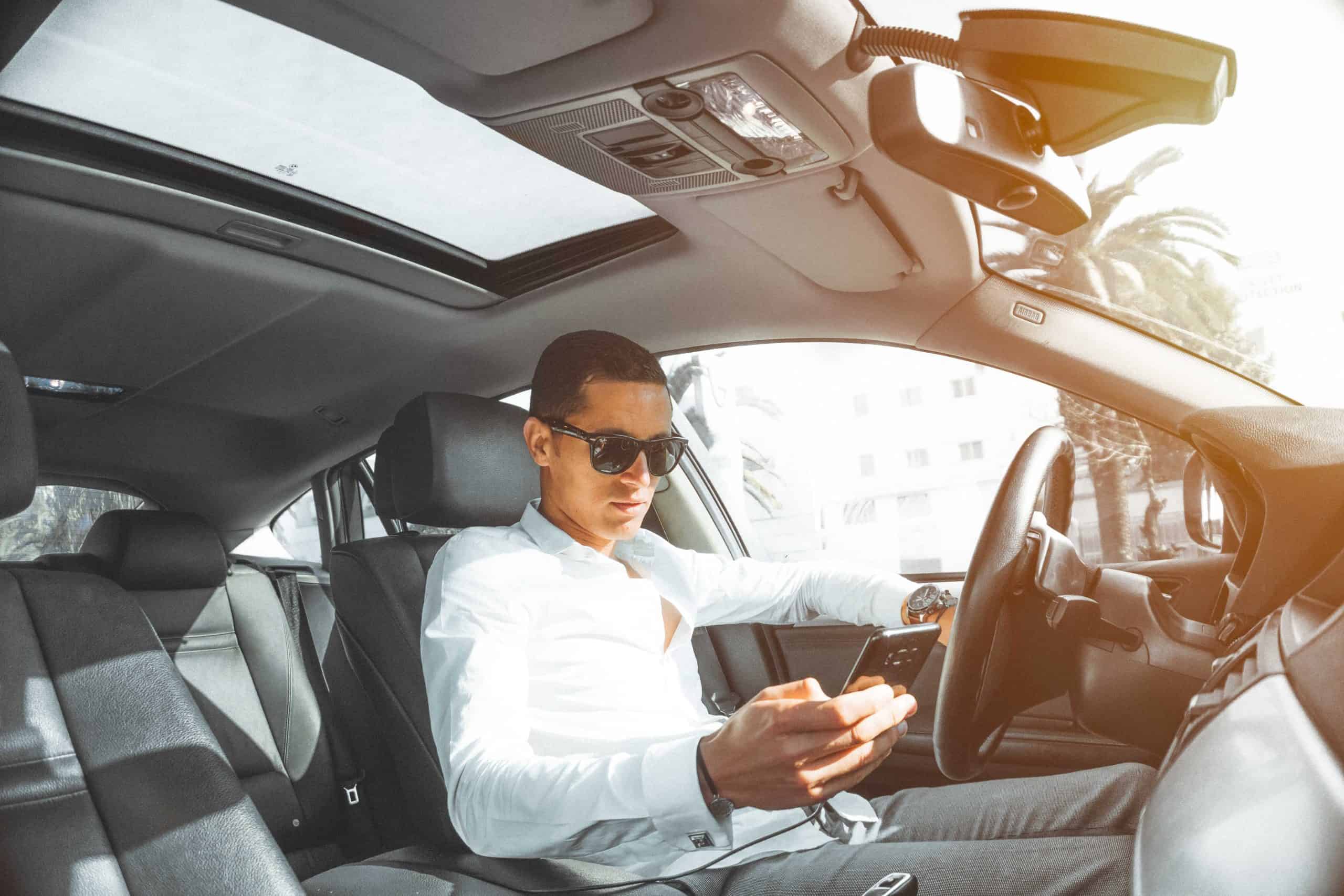 Person in a white shirt and sunglasses is sitting in a car, looking at their smartphone. The car interior includes a sunroof, and sunlight is streaming in.