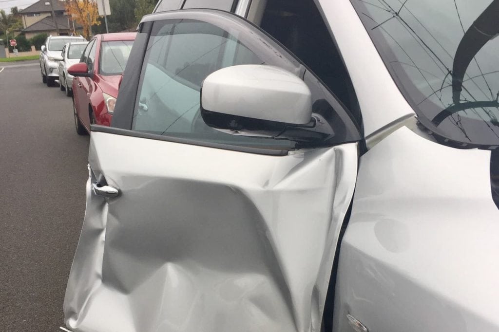 Close-up of a silver car with a large dent in the driver's side door, parked on a street. Other cars are visible in the background.