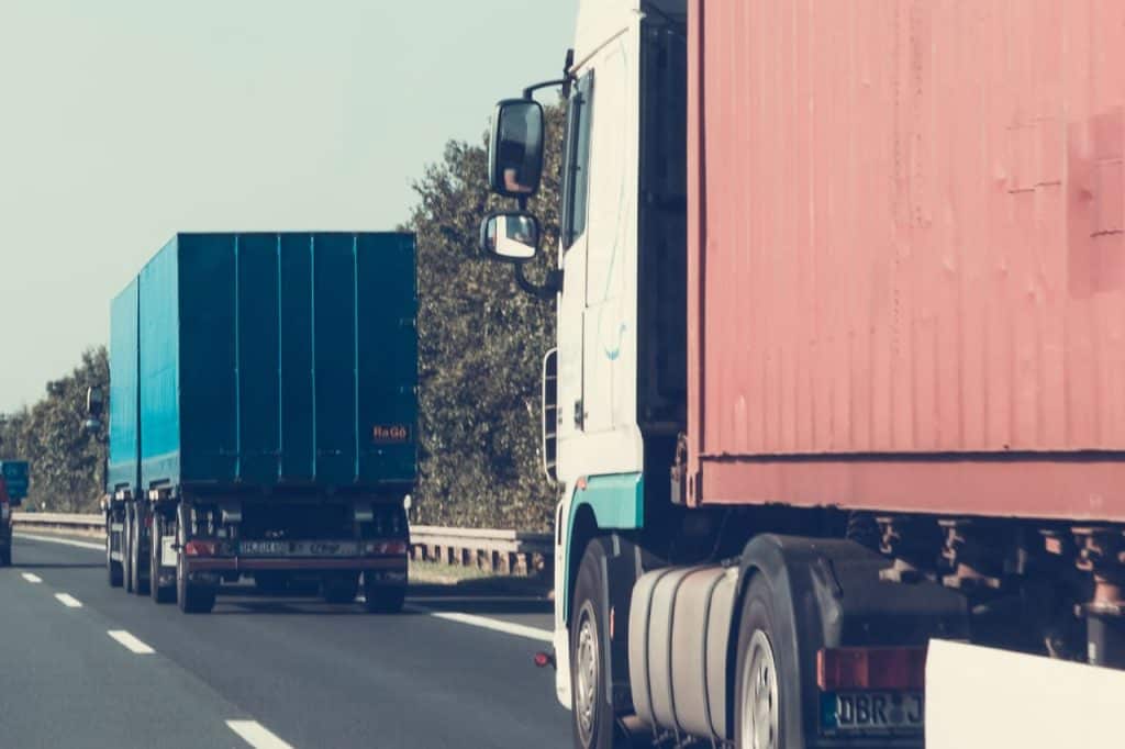 Two trucks drive along a highway, one with a blue freight container and the other with a red container. Trees line the road.