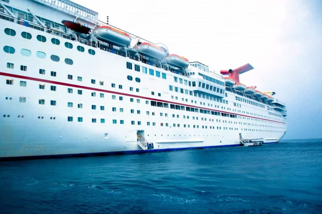 Large white cruise ship with multiple decks and lifeboats, docked at sea. Aqua marine water and overcast sky complete the scene.