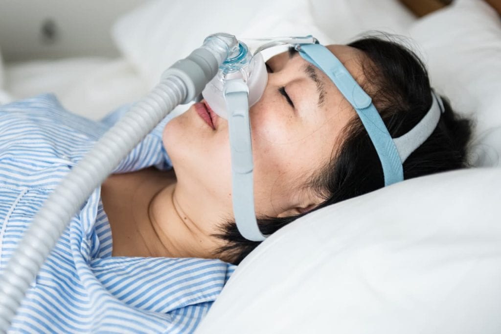 Person sleeping with a CPAP mask on, lying on their back in bed, wearing a blue and white striped shirt.