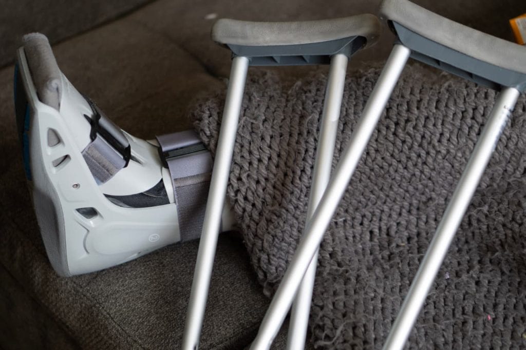 A leg in a medical walking boot rests on a gray knitted blanket, with two crutches placed beside it on a couch.
