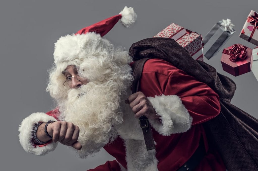 Santa Claus is in motion, carrying a brown sack filled with wrapped gifts. He is wearing traditional red attire, a hat, and checking his wrist as if looking at a watch.