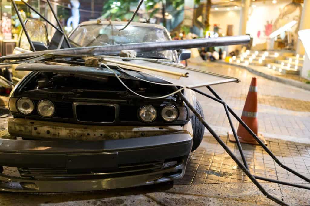 A car with a crumpled hood is covered by fallen metal poles and cables on a city street. An orange traffic cone stands nearby.