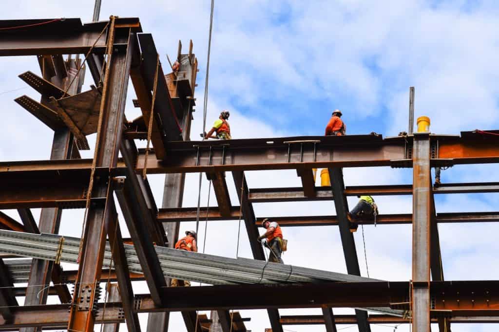 Construction workers in safety gear are assembling a steel framework on a building site against a blue sky. Some are climbing, while others are carrying materials.