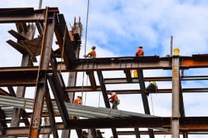 Construction workers in safety gear are assembling a steel framework on a building site against a blue sky. Some are climbing, while others are carrying materials.