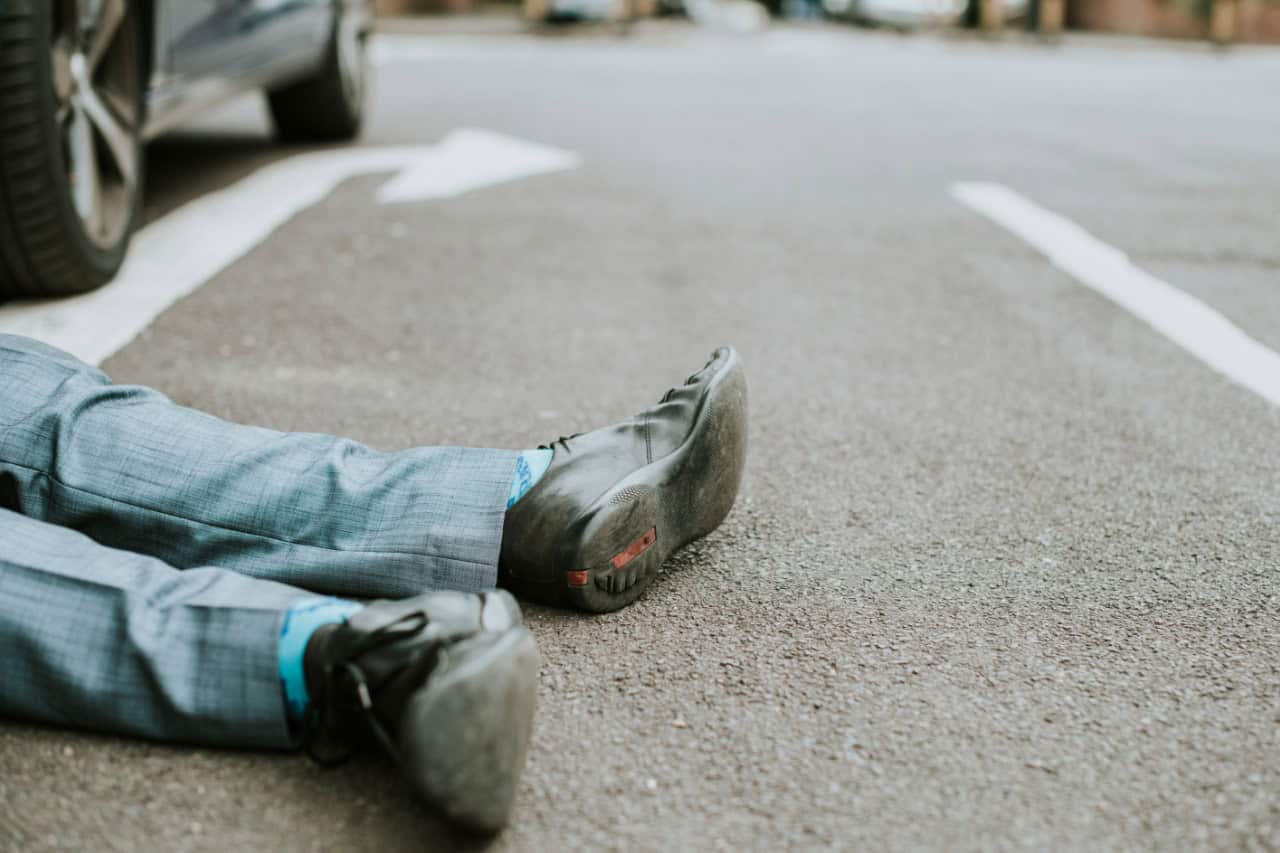 Person in a suit lying on the pavement near a car, with legs visible and the scene focused on their shoes.