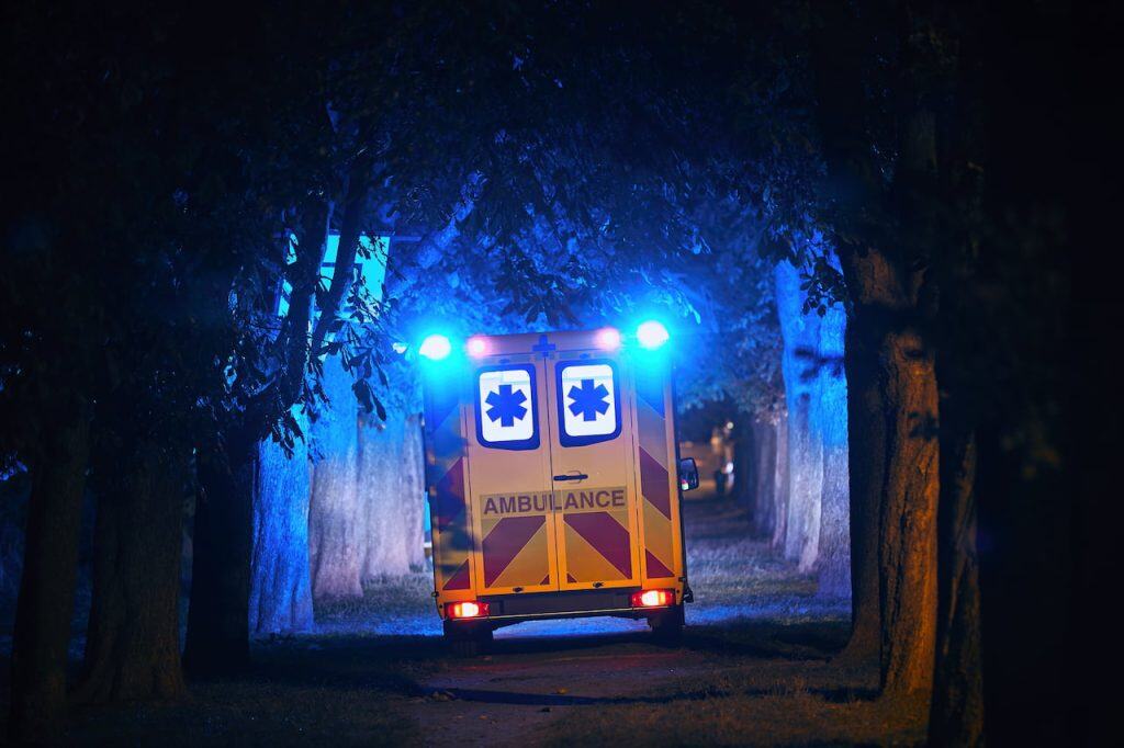 An ambulance with flashing blue lights moves along a tree-lined path at night.