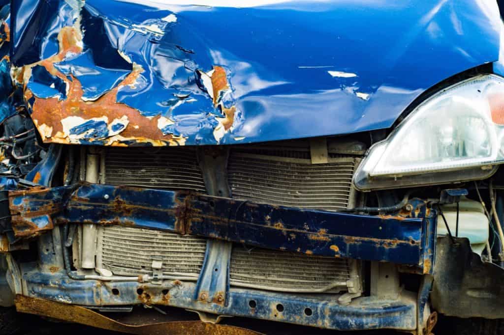 Close-up of a heavily damaged blue car front, with a crumpled hood, exposed radiator, and rusty metal.