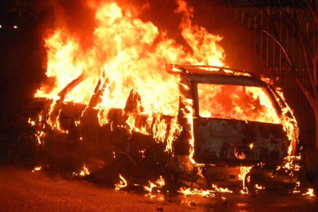 A car fully engulfed in bright, intense flames during a fire at night, with visible damage and no people around.
