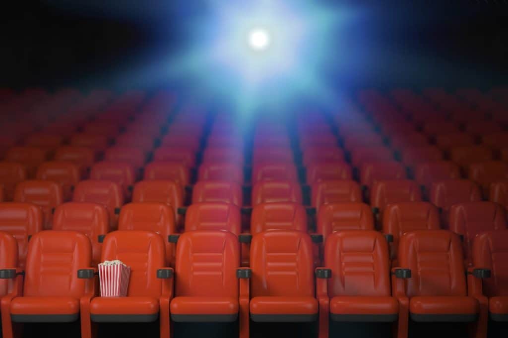 Empty movie theater with rows of red seats and spilled popcorn on one. A bright projecting light illuminates the scene.