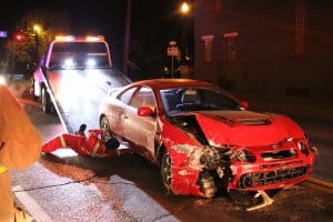 A damaged red car is being loaded onto a tow truck at night. A person in red overalls is guiding the vehicle. The front of the car is severely damaged.