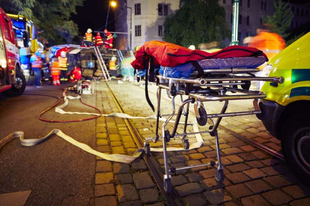 Emergency responders at a nighttime scene with a stretcher, ambulance, and fire brigade. Equipment and hoses are on the ground; responders are actively working.