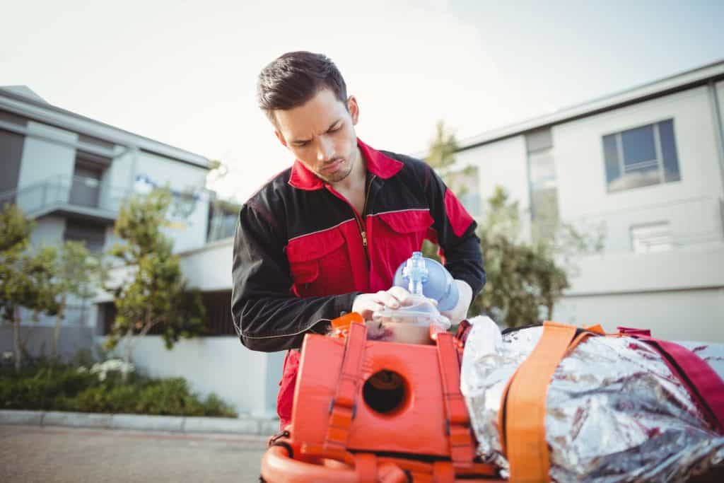 Emergency responder using a bag-valve mask on a person wrapped in a foil blanket on a stretcher, outdoors near buildings.