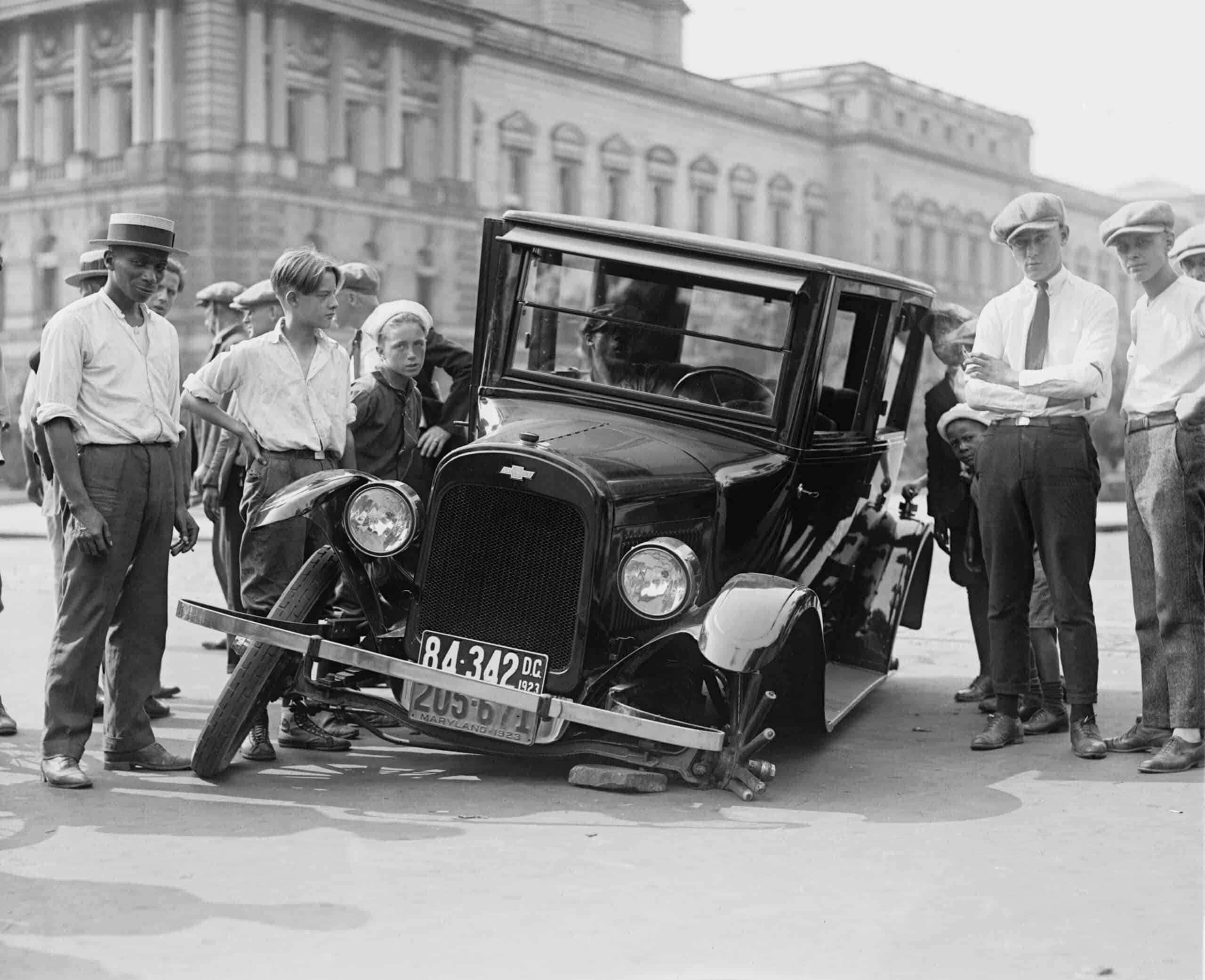 A group of people gather around an old car with a broken wheel in front of a large building.
