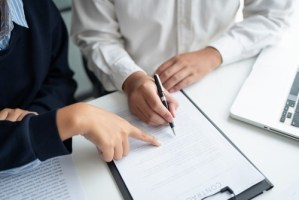 Person signing document on clipboard while another points beside laptop