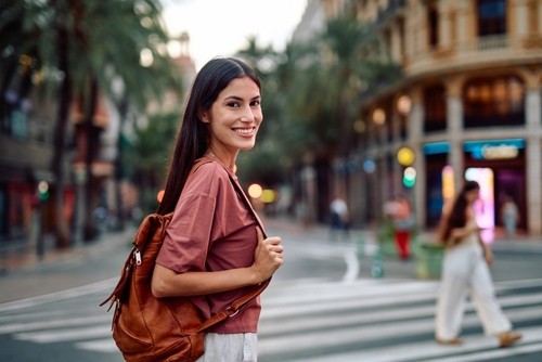 smiling young woman with long dark hair and backpack crossing urban street